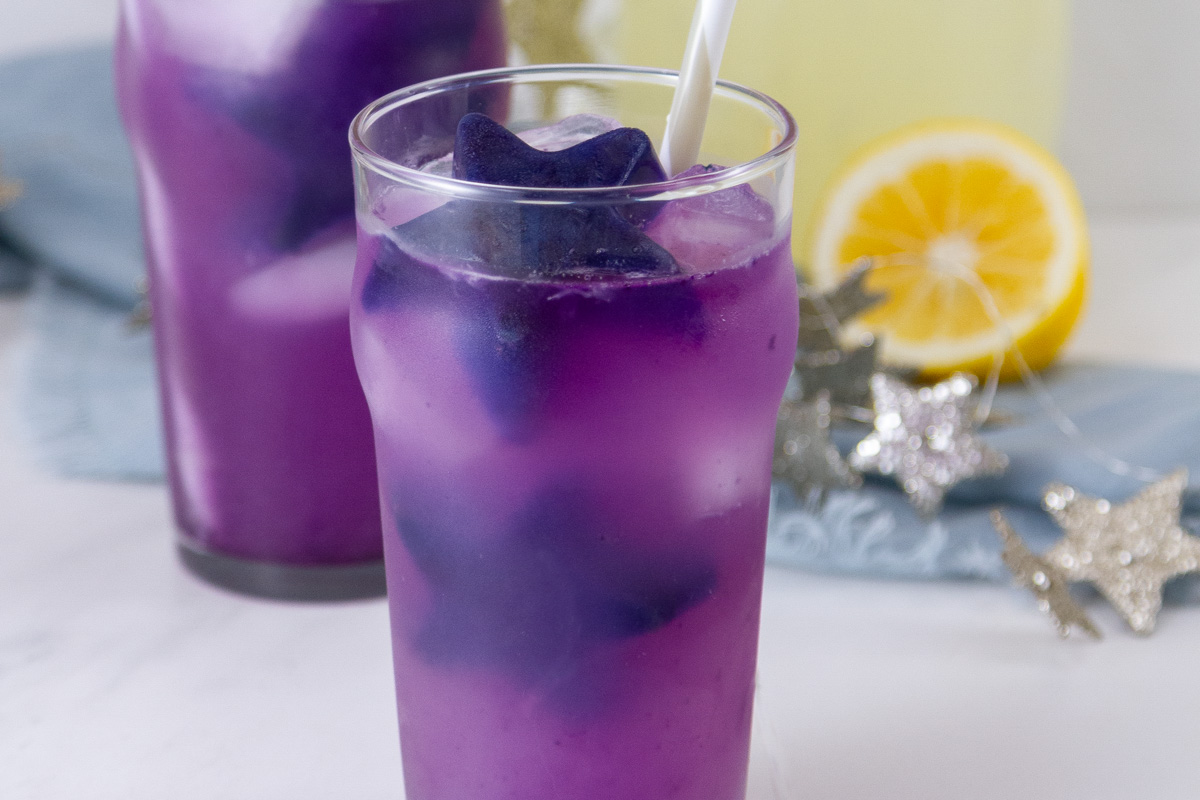 two glasses of galaxy lemonade with purple star ice cubes in them and a lemon in the background.